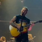 Zach Bryan singing with an acoustic guitar on an arena stage under string lights