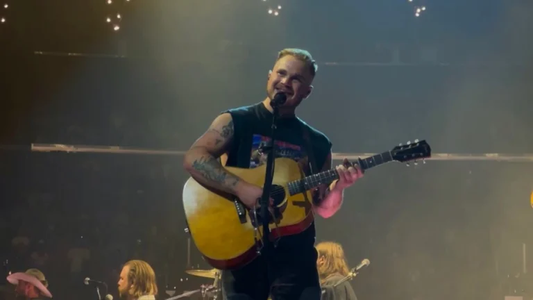 Zach Bryan singing with an acoustic guitar on an arena stage under string lights